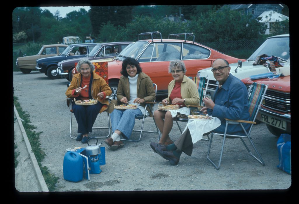An old colour photograph of four people having lunch in front of their car