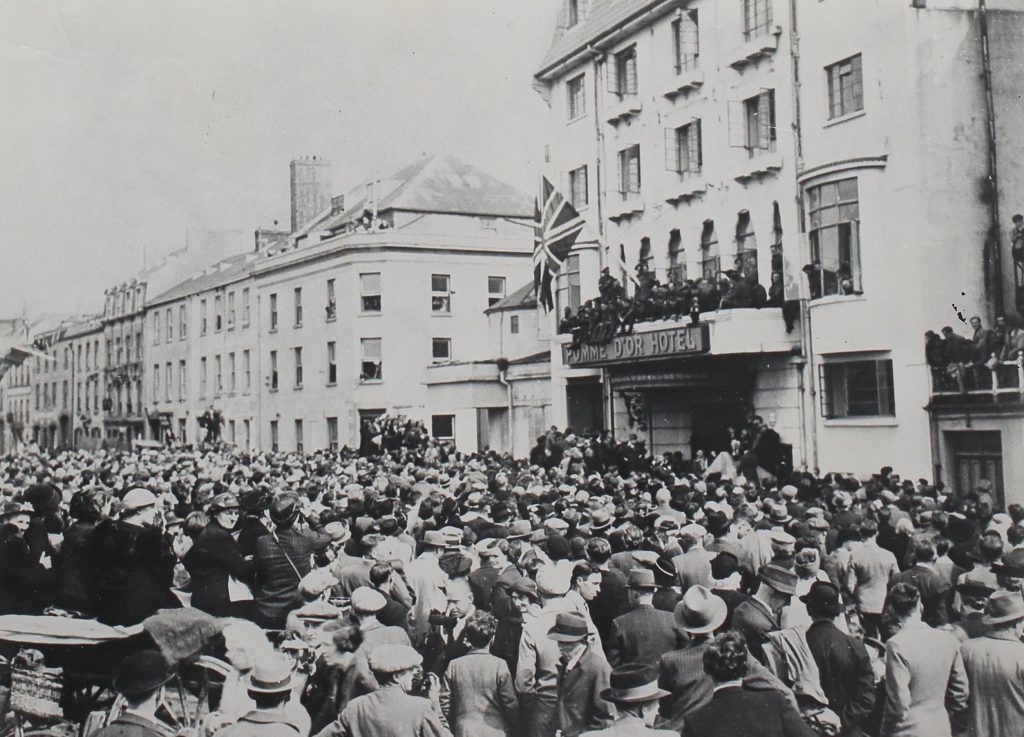 A black and white photograph of a huge crowd filling the street outside the Pomme D'or Hotel