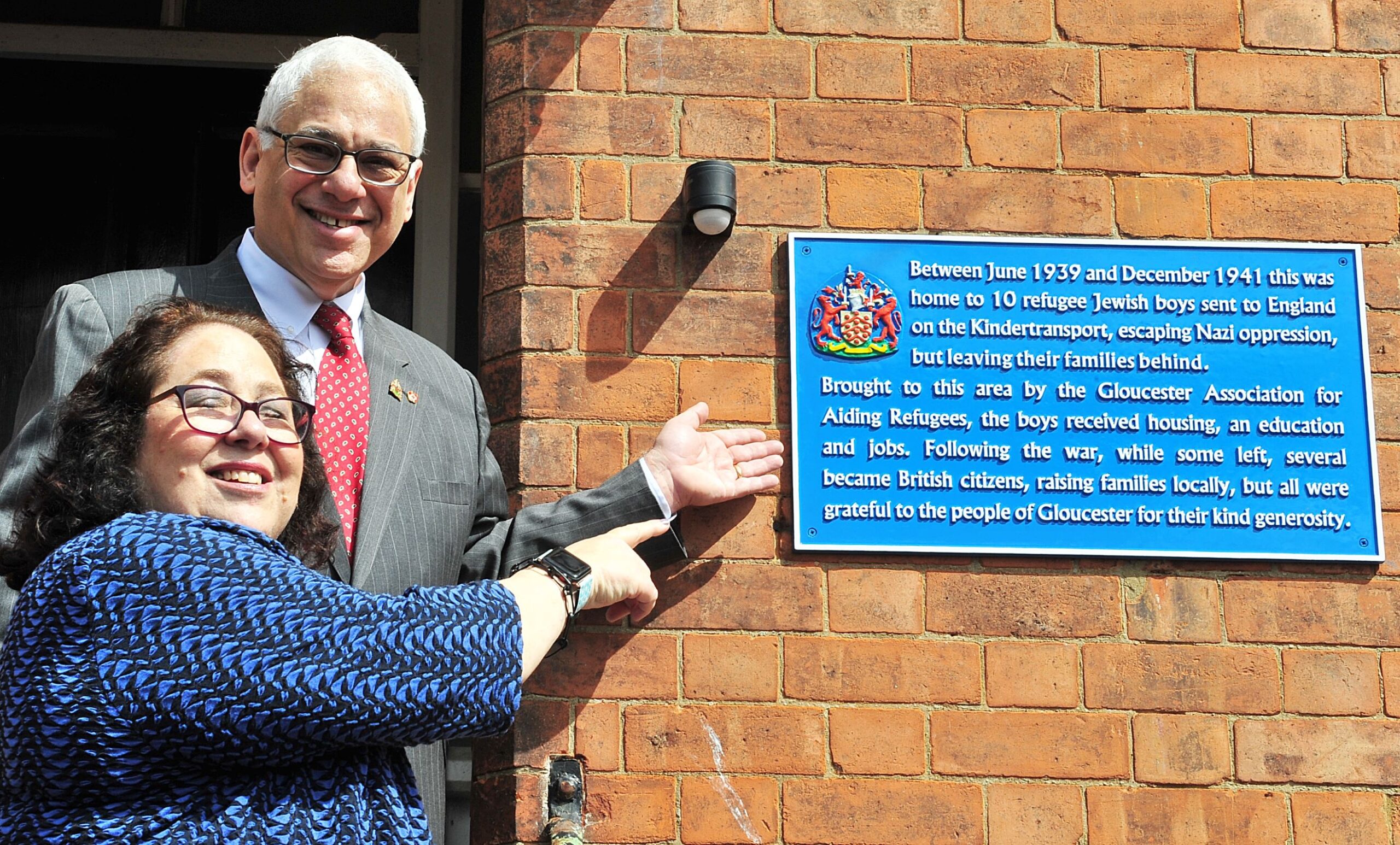 Two people smiling and pointing at a blue plaque on a wall