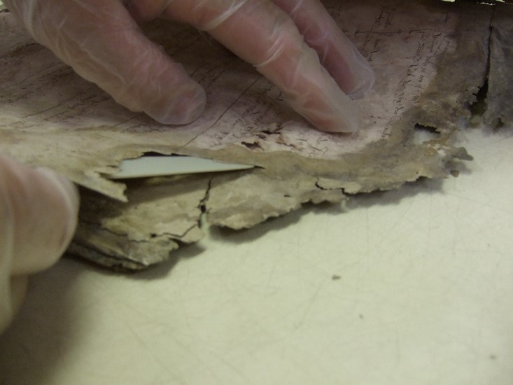 A close-up of the original manuscript that appears in Figure 9. The greyed and deteriorating top edge of the document can be seen, as a gloves hand uses a thin tool to prise two pages apart.