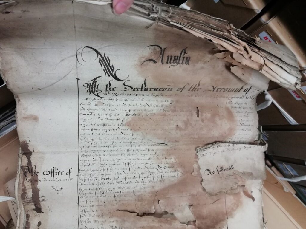An ornately written manuscript roll, displaying crisping at the edges, a rust brown stain down the middle-right of the document, and holes in the paper. A small detached piece of the document can also be seen, placed on top of the roll.