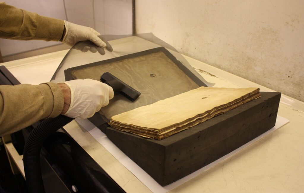 A large manuscript book being treated with a small flat vacuum attachment, which is being applied to the manuscript through a mesh screen.