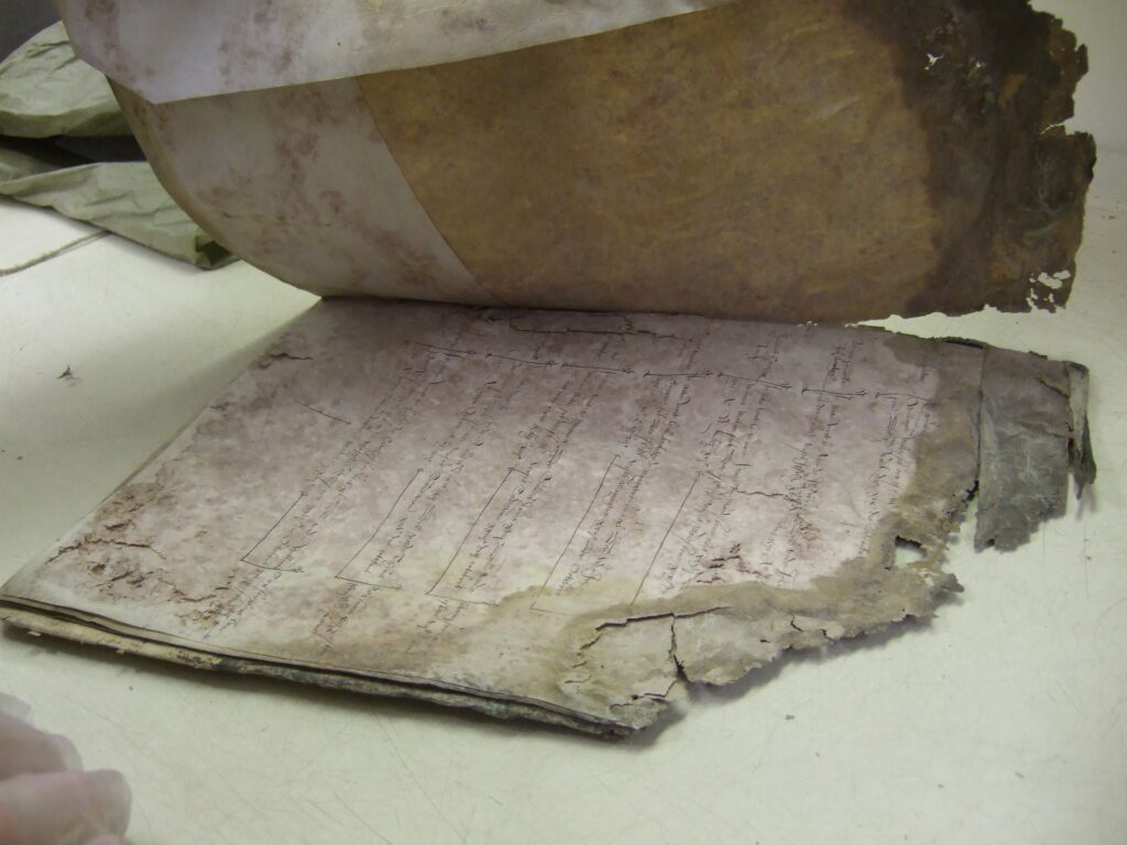 An original paper manuscript, with a page lifted up to show the damage between the pages. They are greyed and deteriorating severely at the edges. Severe discolouration can also be seen.