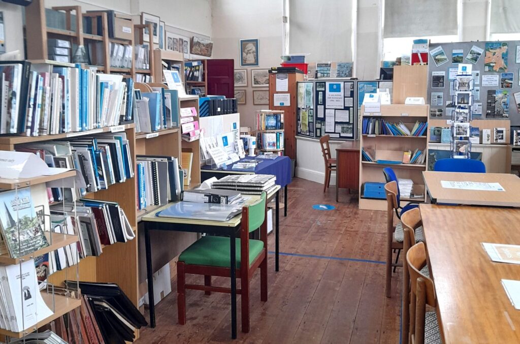 View of the left side of a library space, with a series of shelves on the left filled with books and publications. The room has high, white walls, with windows set into the top of the wall.