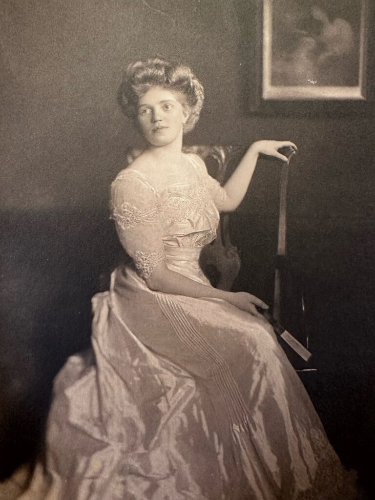 A woman poses for a photo, sitting sideways on a chair with her torso twisted round to face the camera. Her left hand is resting on the top of the chair backrest.