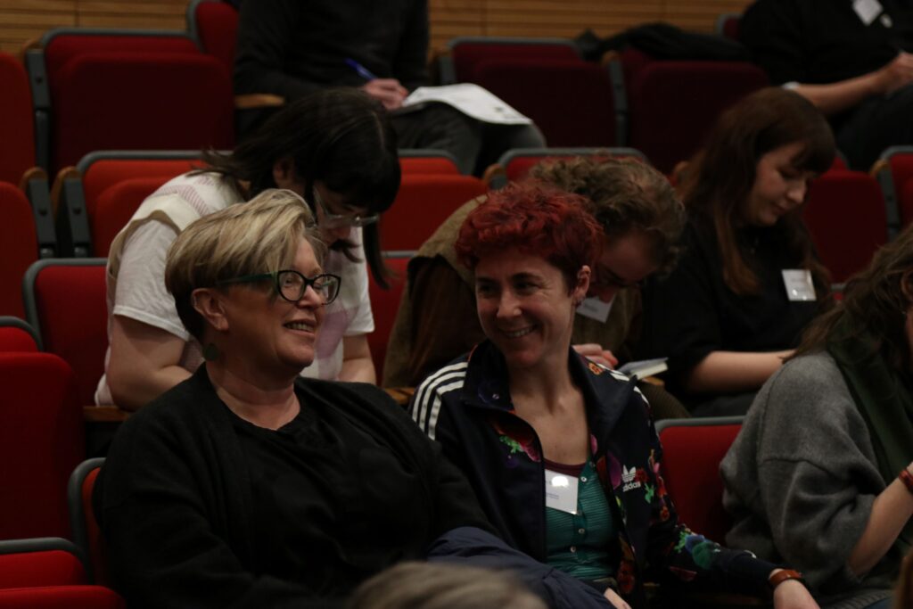 People sat in red auditorium seating, most of them making notes on paper. Two people in the foreground at talking to one another and smiling.