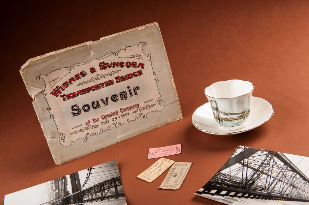 A selection of objects on a terracotta colour table, including a small print that reads 'Widnes & Runcorn Transporter Bridge Souvenir of the Opening Ceremony, May 29th 1905', in a mix of red and black lettering. There is also a small china cup and saucer with an illustration of a bridge on it, two black and white photos of a bridge, and three small ticket stubs.