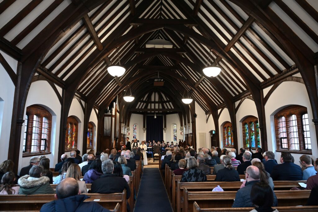 Taken from the rear of a chapel, a number of people are seated in wooden pews and looking towards the altar. A choir is seated at the very front of the chapel, behind four members of the clergy