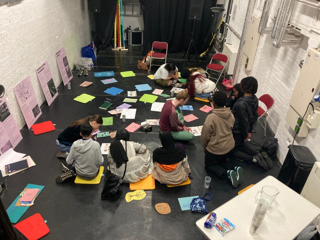 A small group of adults and children sitting or kneeling in a room with white walls and a black floor. On the floor are several pieces of different coloured paper and card, which the group are engaging with