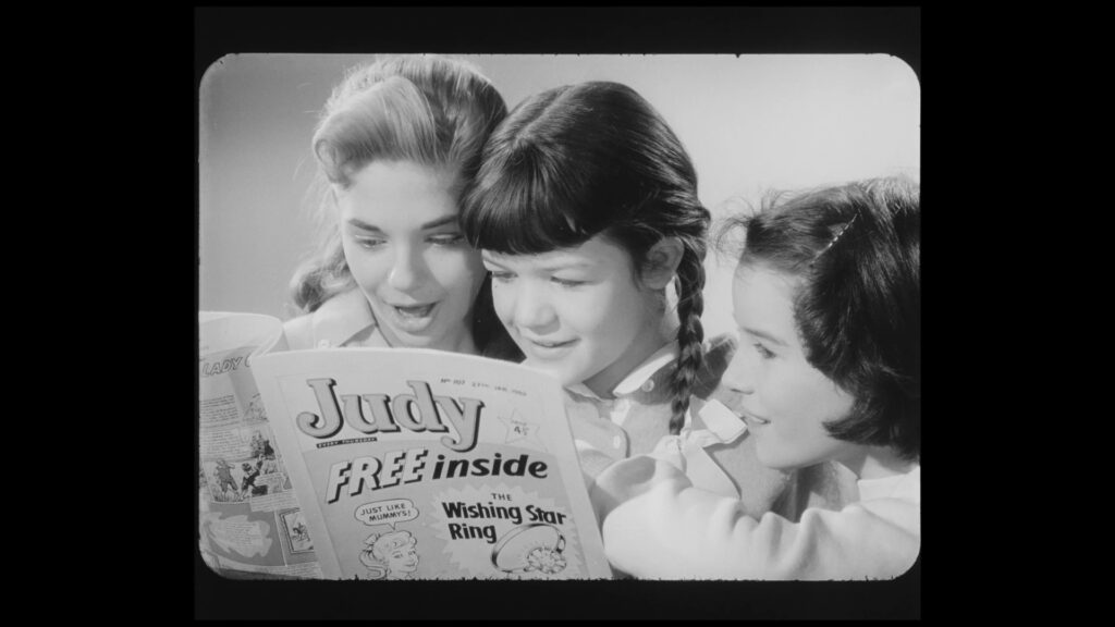 Still from a black and white television advert, in which three girls - one adolescent/young woman, with two younger girls on her left. The adolescent/young woman is holding a copy of 'Judy' comic. The front cover has text reading 'FREE inside - The Wishing Star Ring', with an illustration of a ring with a large stone in it and a girl saying 'Just like mummy's!'