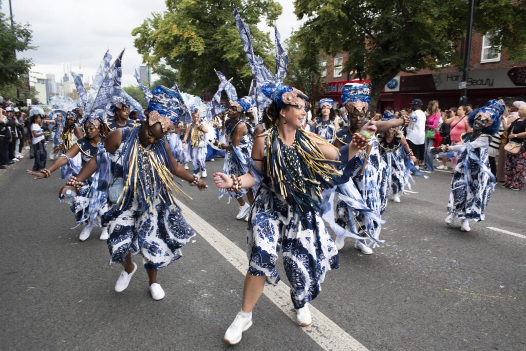 A group of dancers in the middle of a street, dancing in formation. They are wearing matching blue and white costumes and headdresses.