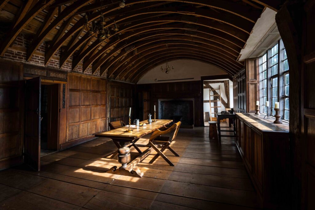 A long wooden-panelled room with a curved ceiling with curved beams. Light is coming through a window on the right, illuminating a table and two chairs in the middle of the room.