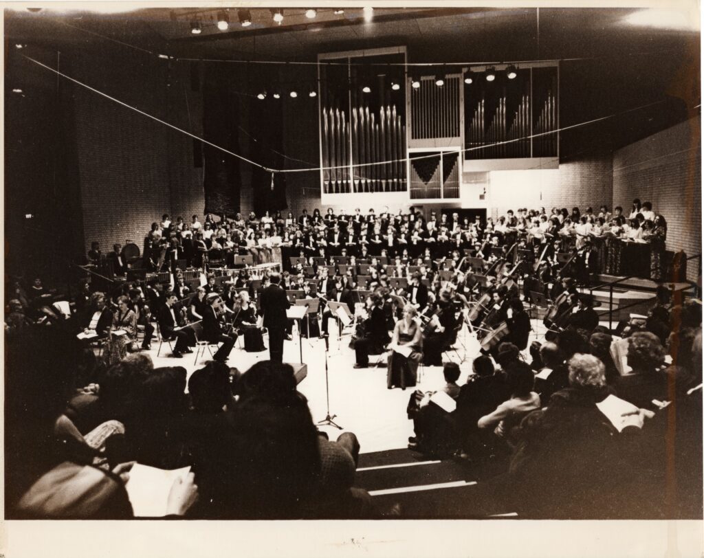 Black and white photo of a very large orchestra, in formal attire. A few audience members are in the foreground.