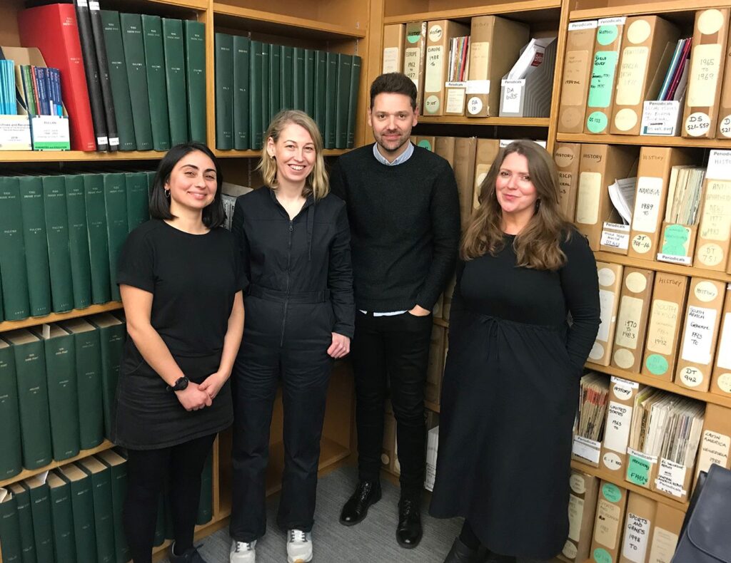 Four people standing in the corner of a room, posing for a photo. They are all wearing black. Behind them are a selection of large green bound documents on one wall, and a series of cardboard archive boxes on the other wall