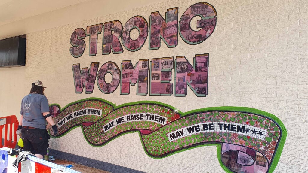 Mosaic mural on a white brick wall - 'Strong women' in pink letters with green detailing, and a flowing green and pink scroll below it that reads 'May we know them, may we raise them, may we be them'.