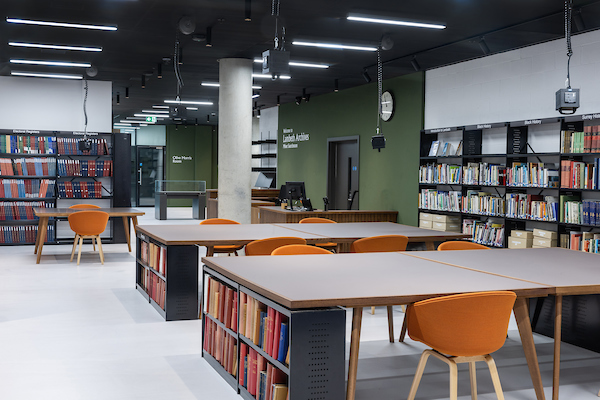 View of a reading room - the right-hand wall and the far wall are full of books on bookshelves, while the middle of the room has two tables with room enough for four orange chairs at each