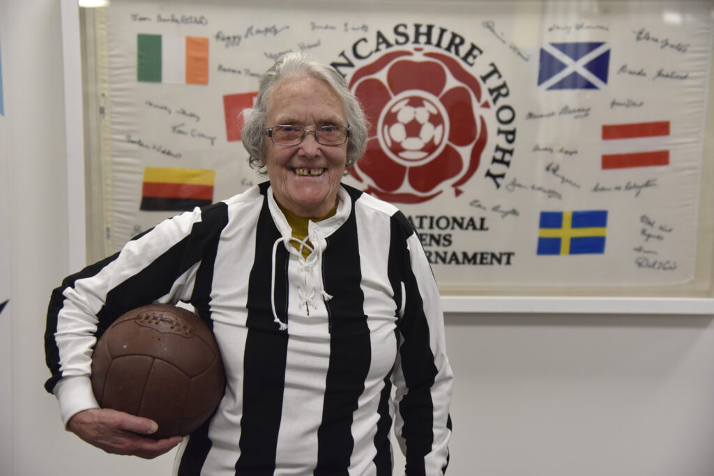 An elderly woman posing for a photo, wearing a retro black and white striped football shirt, with a vintage-style football under her right arm.