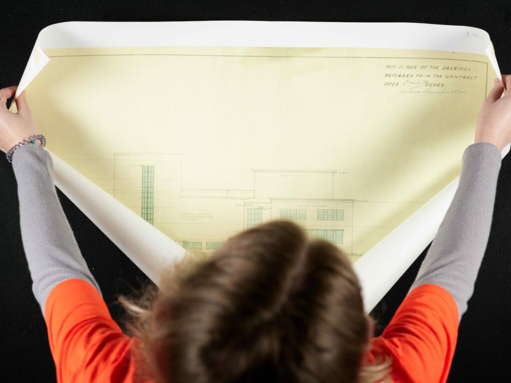 Birds-eye view of a person unfurling a plan drawing of a building, on a large black cloth. The person has brown hair, and is wearing a grey long-sleeve tee with an orange t-shirt over the top.