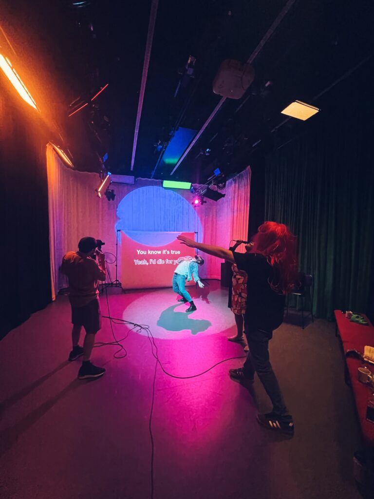 Four people stood in a studio environment, with strong pink lighting and green floor-to-ceiling curtains surrounding the space. Three have microphones and are singing along to words being projected onto a screen, while the other is dancing under a spotlight.