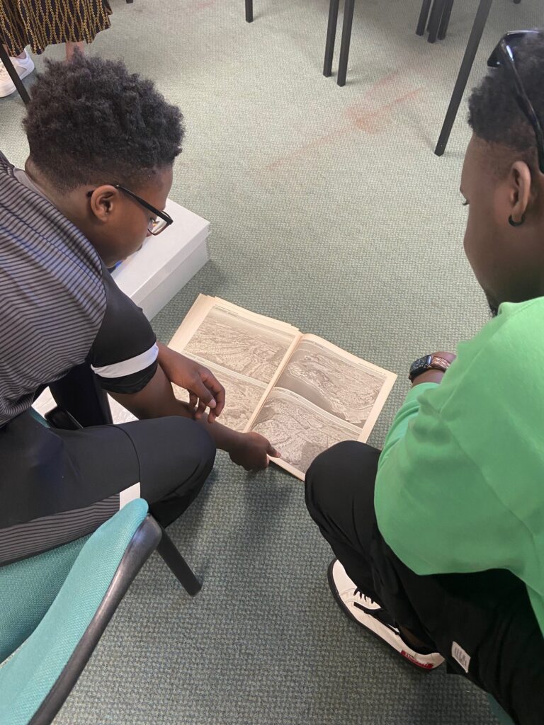 Two boys sat on chairs lean forward, looking down at a book that is resting on the green-grey carpet floor. The book contains black and white aerial photographs, and the boy on the left is reaching with his right arm to turn the page.