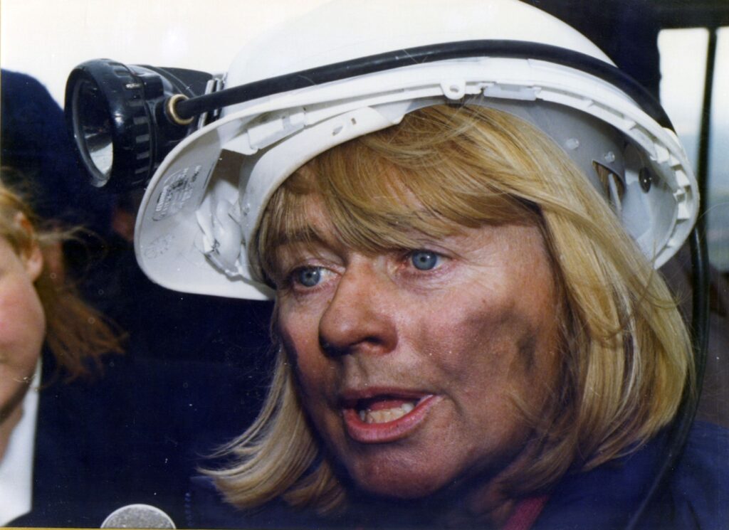 A close-up photo of a woman wearing a white hard hat, which has a black torch strapped round it with a thick black cable. The woman has shoulder-length blonde hair and blue eyes, with coal marks on her face, and her mouth is open as if she is speaking to someone.