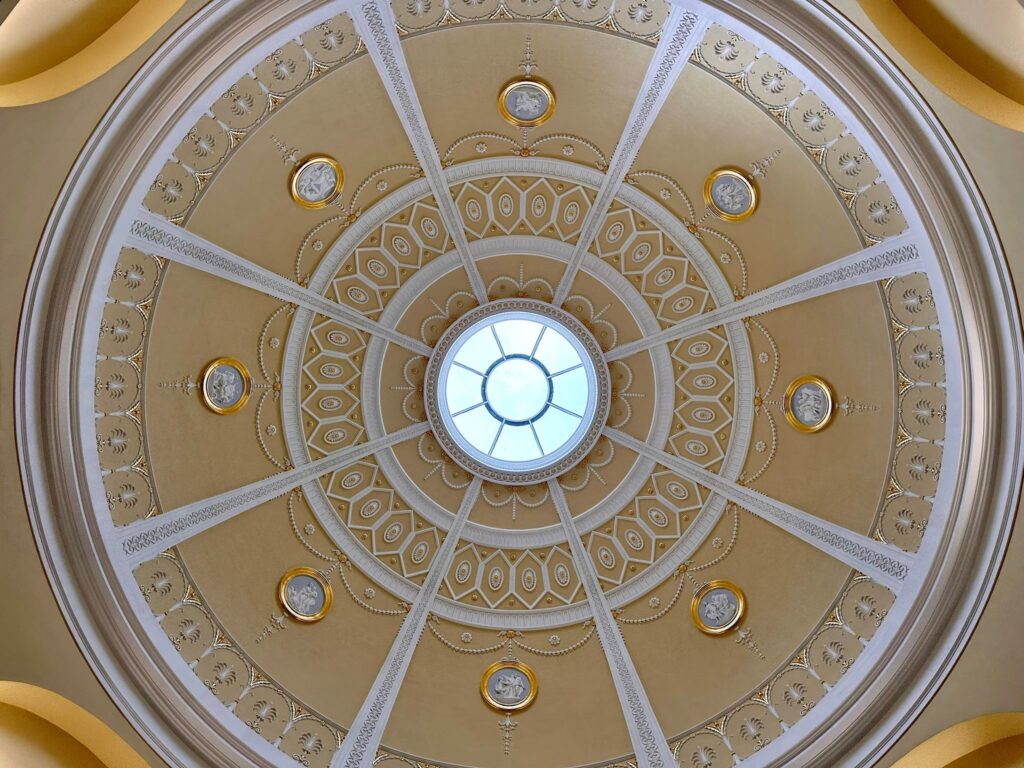 A domed area of ceiling, as viewed from below. There is a small circular glass window in the centre, with a series of decorative plaster and gold-coloured features centred around it.