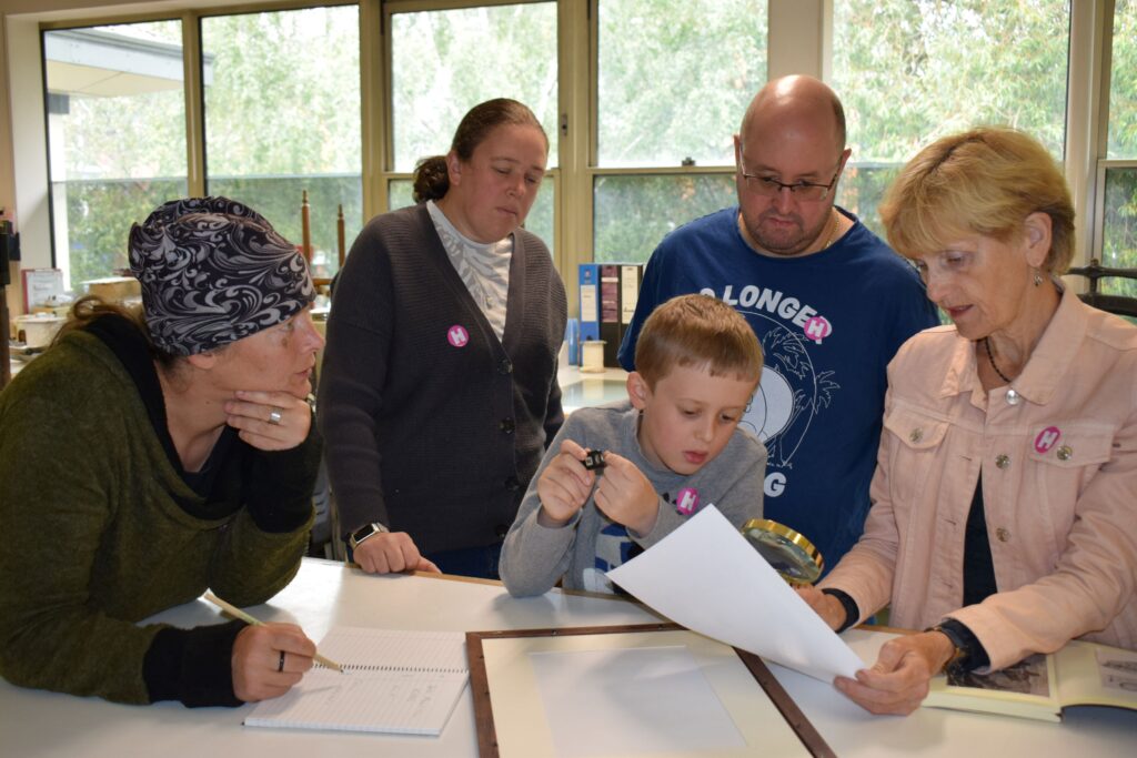 A group of four adults and one boy crowded round a table, looking at a piece of paper that is being held by the person on the right of the photo. They are holding the piece of paper in their left hand, and a magnifying glass in their right hand.