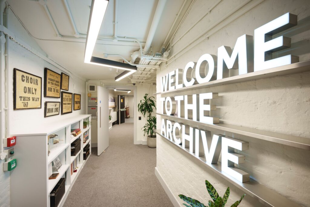 A white-painted corridor that has 'Welcome to the archive' on the right-hand wall, in large illuminated letters. Some letterpress prints are on the left-hand wall, such as 'Choir only this way [with a hand pointing to the left]'