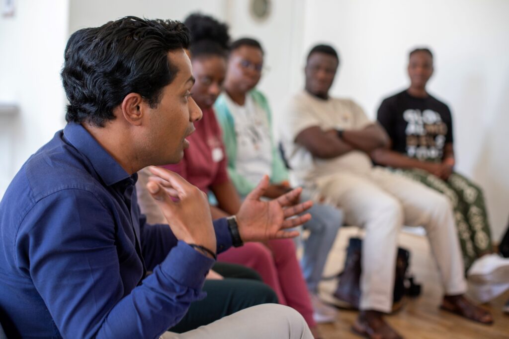 Six people sitting in a semi-circle, each one sat a little further from the camera. The person closest to the camera is talking animatedly, and the rest of the group are looking at him.