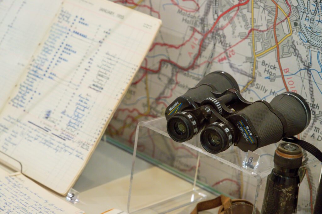 Part of an exhibition display - a pair of binoculars are resting on a clear acrylic shelf, with a large print of a section of ordnance survey map behind it. To the left of the binoculars is an A4 lined booklet, propped up and open, with the pages written in with blue ink.