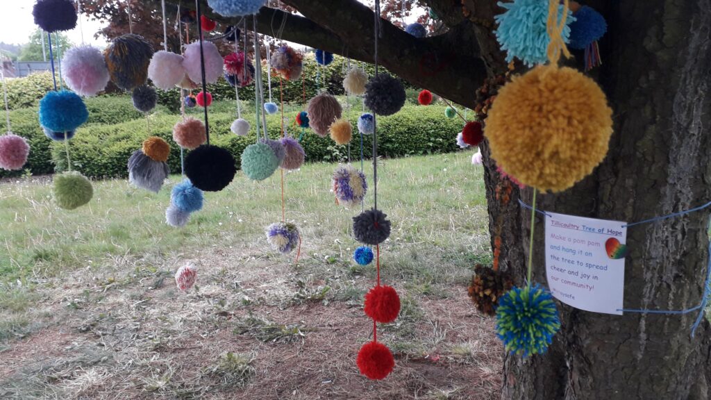 A large tree trunk is on the right, with a couple of thick branches coming from it. A large number of differently-coloured pom poms are hanging from the tree, and tied round the tree is a printed message: 'Tillicountry Tree of Hope - Make a pom pom and hang it on the tree to spread cheer and joy in our community! #staysafe'