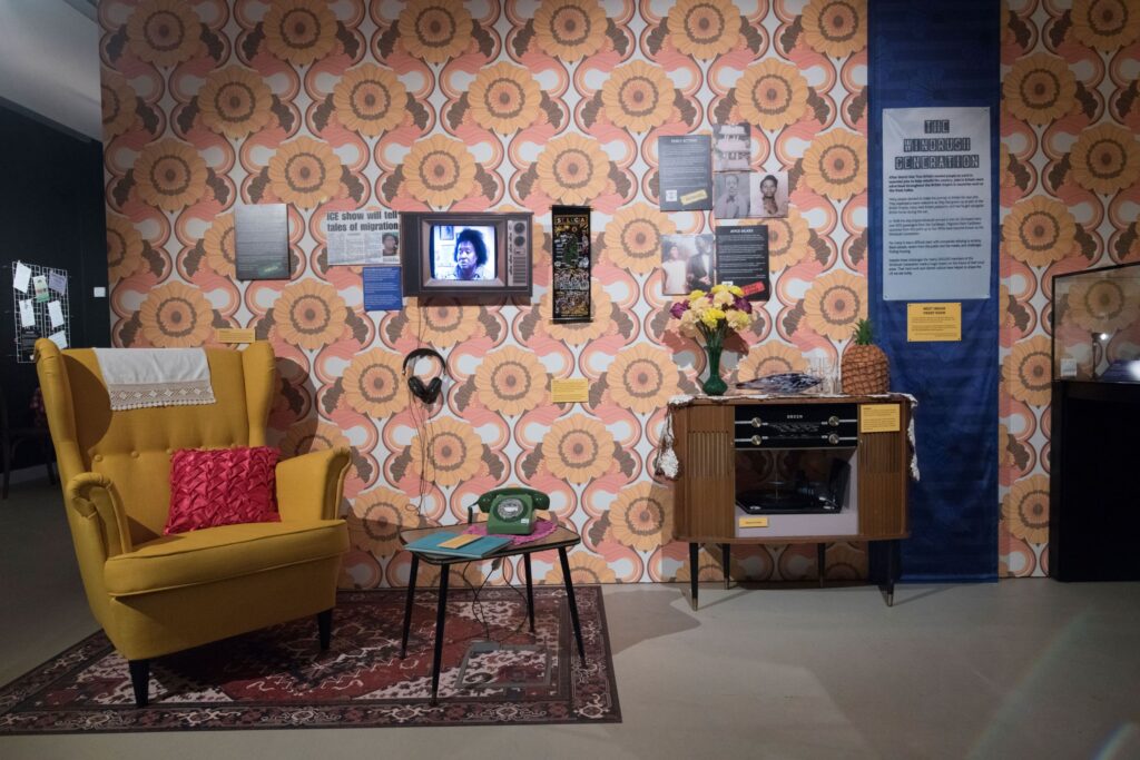 An exhibition space featuring a replica living room with colourful, stylised sunflower wallpaper, a yellow wingback chair, a small replica television in the wall, a vintage record player, and a green telephone on a side table. Text on the wall is titled 'The Windrush Generation'.