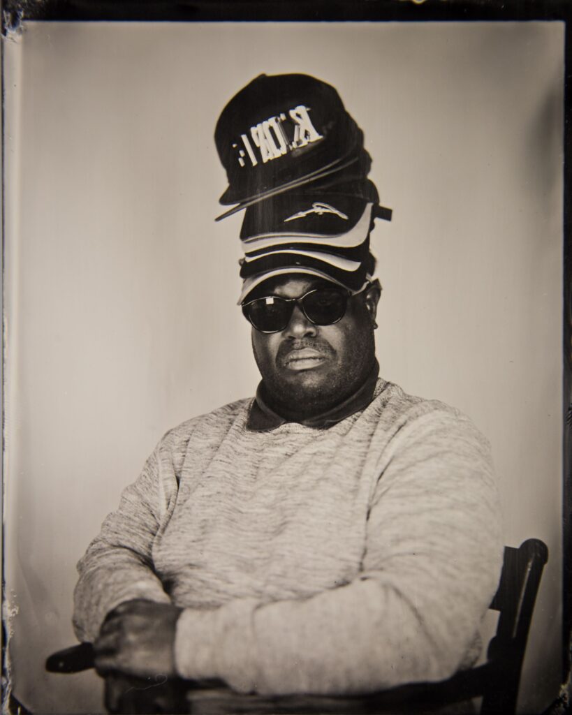 A black and white photo of a man in a light-coloured sweater, with a dark-coloured polo shirt collar visible, sitting in a wooden chair. He is wearing a pair of large black sunglasses, and has several caps stacked atop one another balanced on his head.