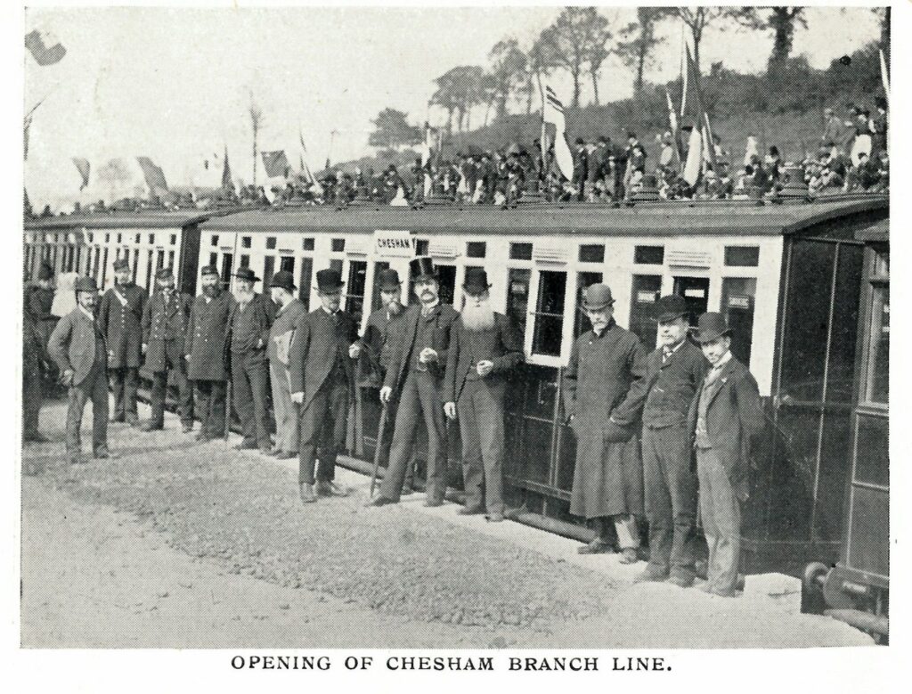 Black and white image displaying a number of men in contemporary dress - top hats, bowler hats, suits and long coats, most of them with facial hair - standing in front of a train carriage. A number of spectators are behind the carriage, on a hill. The title of the image is 'Opening of Chesham Branch Line.'