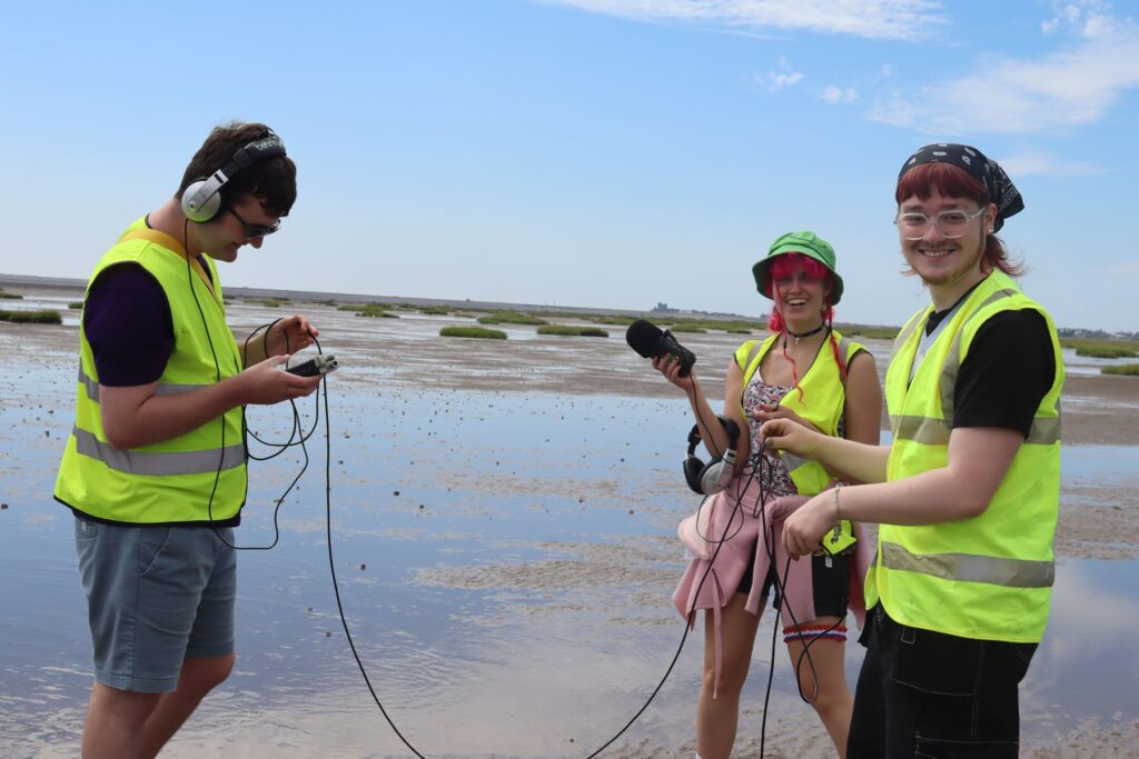 Three people wearing high-vis jackets stood on a flat, wide beach. Two are stood together on the right, looking at the camera. Between them they are holding elements of audio recording equipment such as cables, headphones and a microphone.