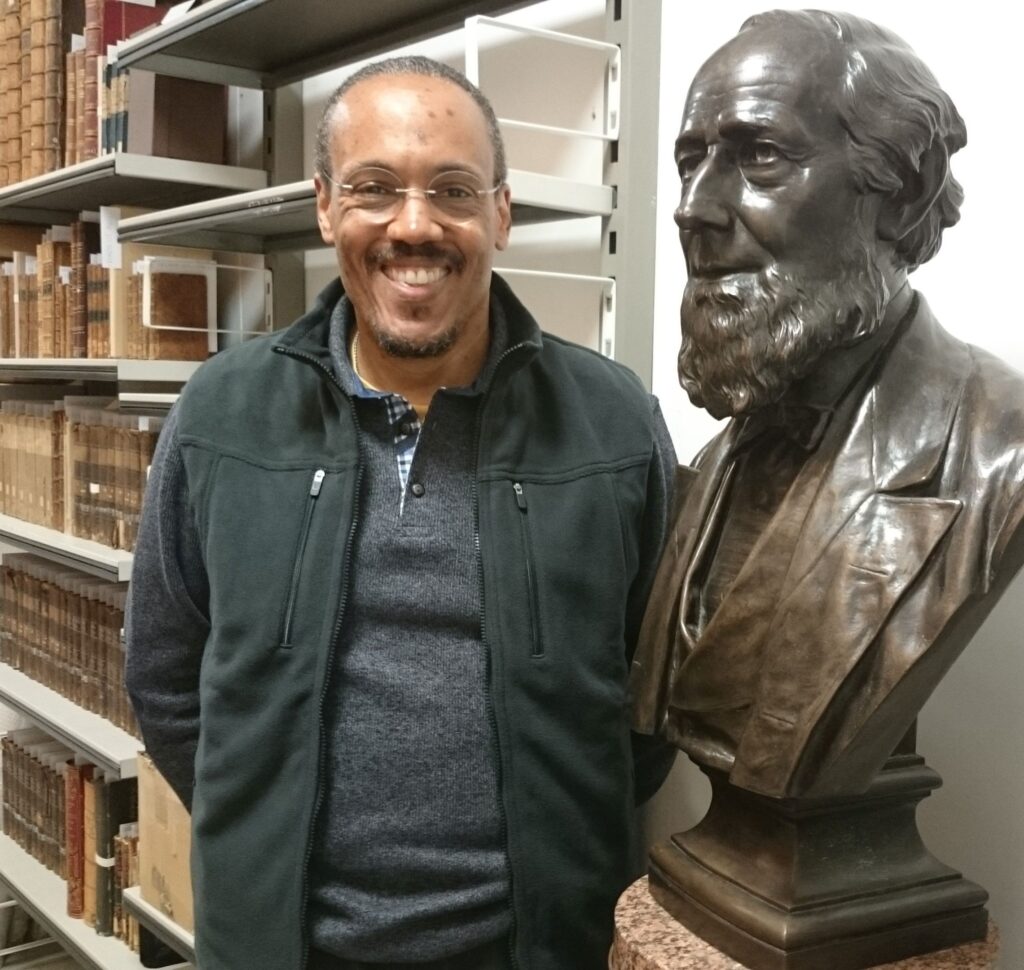 A man stands, smiling, posing for a photo next to a bronze bust of a bearded man. The man posing for the photo is holding his hands behind his back, and several hardback-bound documents are on shelves behind him.