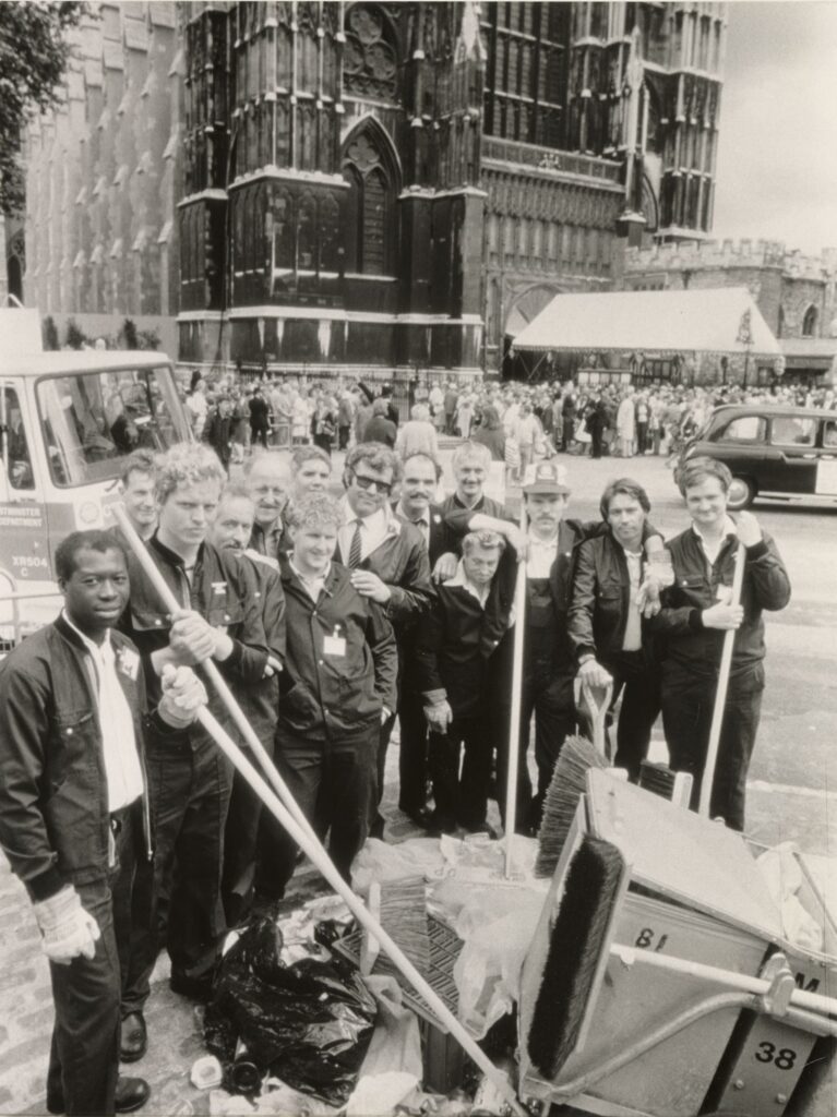 Black and white image of 14 men posing for a photo, some holding brooms. There is a pile of rubbish in front of them and, behind them, Westminster Abbey, with a crowd gathered outside.