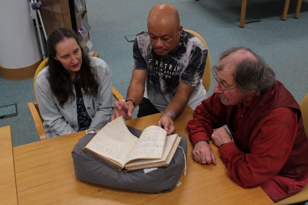 Three adult people at around the corner of a table, all looking at a bound document that is resting on a cushion on the table. The person in the middle is handling the document, and is in the process of turning a page.