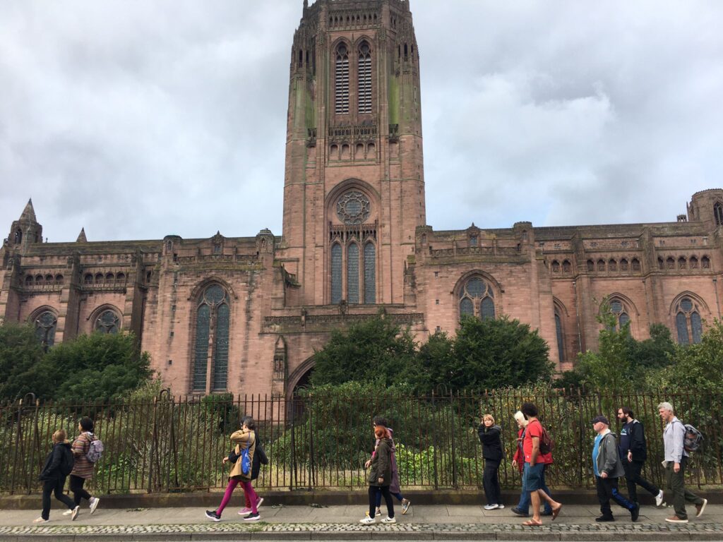 A large red brick church, with a central spire rising from it. Twelve people are walking past on the pavement in the foreground, in a loose group.