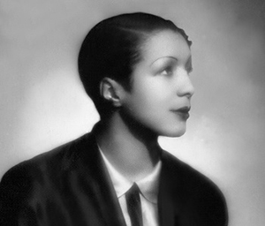 A black and white photo of Evelyn Dove; Evelyn is sitting, her face shown in profile as she faces left. She is wearing a suit and tie, and has a short hairstyle.